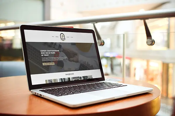 A silver laptop on a wooden table displaying the home page of TM & KM Construction LLC. The website features a header with the company logo and a hero image of two people shaking hands over blueprints.